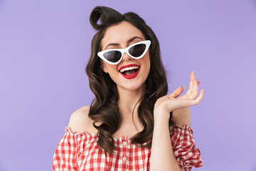 Portrait of joyful pin-up woman 20s in american style wear and retro sunglasses smiling at camera