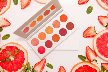 Cute flat lay with palette of cosmetics with fresh fruit, cut strawberries and grapefruit or red orange, mint leaves