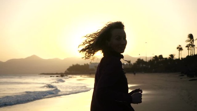 Slow Motion Close up silhouette of Young Woman jogging on a beach with hair blowing in wind looking at sunset over ocean.