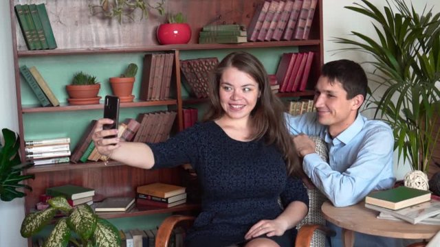 The guy and the girl are sitting in the home library, a man in a blue shirt and jeans, a girl in a dark dress. A girl and a guy pose, make a selfie.