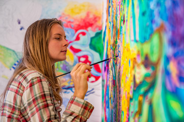 Joyful young female artist painting on the wall, using brush and bright acrylic paints - obrazy, fototapety, plakaty