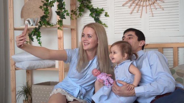 Smiling family in bed, where dad and mom play with their little daughter, mom takes a selfie of her family, takes pictures. Smiles, hugs, happy family.