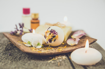 Various spa bath products on brown wooden tray creamy bath bomb, soap bar with cotton string,flower petals, spa candles lit, essential bath and massage oils in background. Instagram filter style.