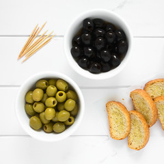 Pitted black and green olives in bowls with crostini on the side, photographed overhead on white wood with natural light