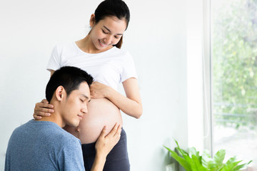 Happy Asian man is listening to his beautiful pregnant wife's tummy and smiling.