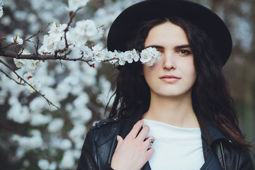 Outdoor spring portrait of fashion sensual young girl posing in amazing blooming garden. Wearing a hat and leather jacket, feminine look, elegant glamorous style. On the background of cherry blossoms