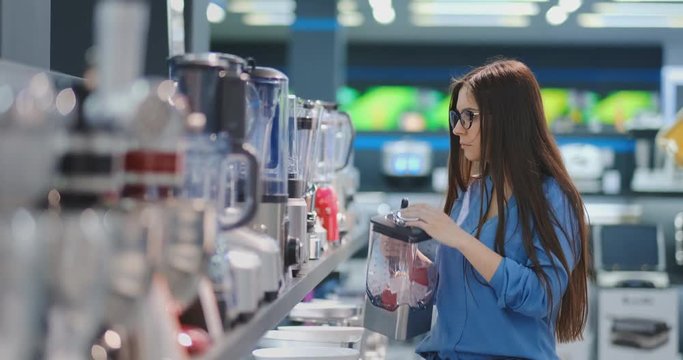 Young beautiful woman in the appliances store is choosing for its kitchen a blender looking and holding in her hands various models.