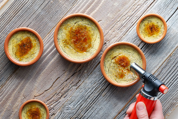 Making creme brulee. Flat lay on rustic wooden table with ceramic brown dishes and flaming torch in female hand