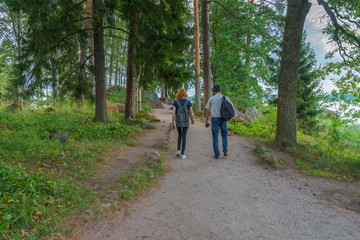 Dark-haired middle-aged man and red-haired young lady walk along forest road. Tourists on the beautiful landscape background. Monrepos Park, Vyborg, Russia. Travel concept