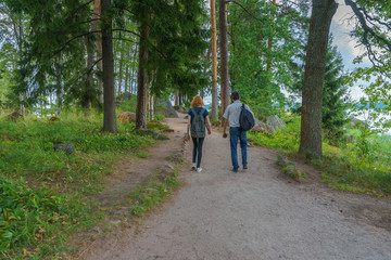 Fototapeta premium Dark-haired middle-aged man and red-haired young lady walk along forest road. Tourists on the beautiful landscape background. Monrepos Park, Vyborg, Russia. Travel concept