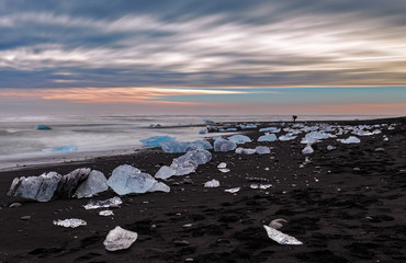 Ice rocks pushed out of the ocean onto a black beach in Iceland with a photographer in the background.