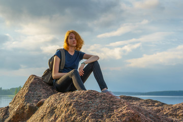 Naklejka na ściany i meble Pretty young lady with red hair sitting on stone near bay shore in summer evening. Tourist on the beautiful landscape background. Monrepos Park, Vyborg, Russia. Travel concept