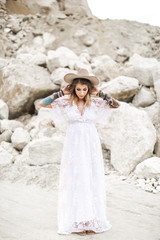 A pretty young woman bride in light brown hat and boho dress is standing on the background of desert canyon. Stylish young girl in long white dress has tattoos on the hands