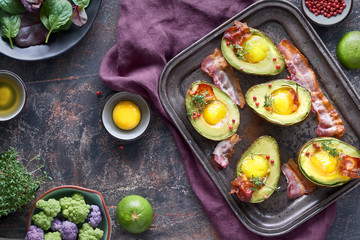 Baked avocado with egg and bacon on a metal baking tray, flat lay with ingredients and herbs on dark