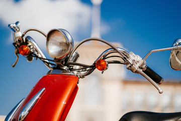 Closeup chrome detail and headlamp of orange retro vintage scooter under blue sky and sun in front of defocused tower