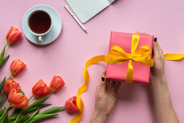 Female hands holding red gift box with yellow ribbon on pink background. Background with coffee, laptop and flowers