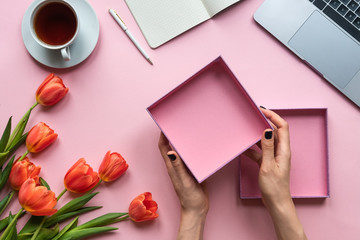 Female hands holding open empty box on pink background. Background with cup of tea, laptop and flowers