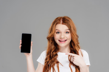 Naklejka premium beautiful redhead girl pointing with finger at smartphone with blank screen, looking at camera and smiling isolated on grey