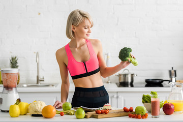 attractive blonde girl in sportswear holding green broccoli in kitchen