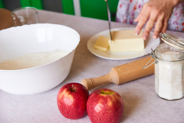 Woman cutting butter on white plate for homemade pie on white tabletop with apples, rolling pin and a bowl of flour