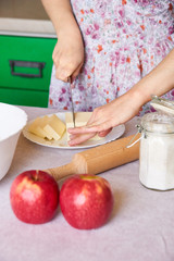 Woman cutting butter on white plate for homemade pie on white tabletop with apples, rolling pin and a bowl of flour