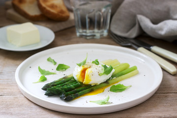 Breakfast consisting of poached egg and boiled asparagus with butter and lettuce on a wooden surface. Rustic style.