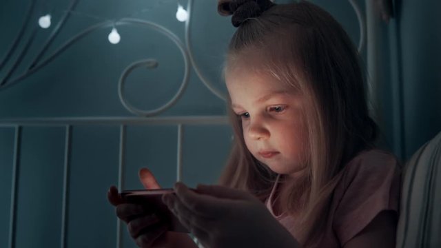 Little girl is looking at the phone. The child is playing on the smartphone. A girl uses the phone in the dark before going to bed. 4k