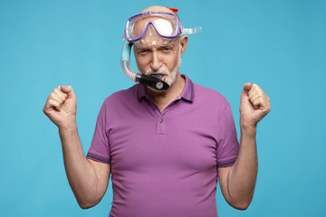 Senior people, retirement, summertime and holidays concept. Funny elderly mature bald man wearing violet t-shirt, snorkeling mask and tube posing at blue blank studio wall, clenching his fists