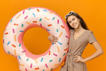 Horizontal image of carefree joyful young Caucasian girl learning how to swim, going to beach, carrying colorful inflatable swimming ring, holding hand on her waist and smiling happily at camera