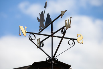 A weather vane attached to the roof of a house facing west with blue skies behind