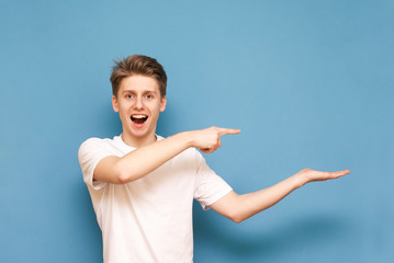 Happy young man in a white T-shirt stands on a blue background and shows with his hands and an empty mission for advertising, smiles and looks at the camera. Isolated. Copyspace