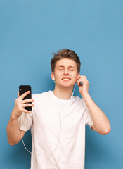 Handsome young man listens to music in his headphones with his eyes closed,holding a smartphone in his hands and enjoying a blue background.Guy listens to his favorite music in the headphones.Isolated