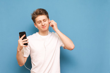 Satisfied teenager wearing a white T-shirt listens to music in his headphones with his eyes closed and holds a smartphone in his hands, isolated on a blue background. Copyspace