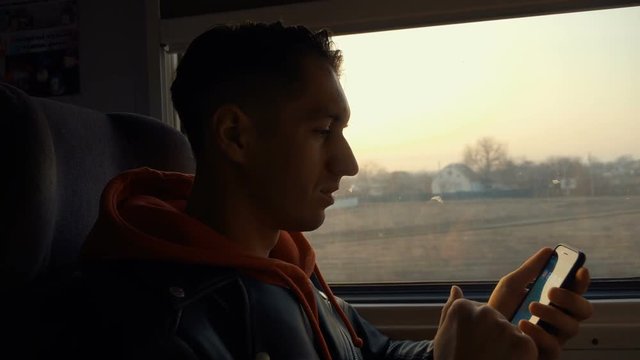 Happy, young man using smartphone during train ride and sitting opposite the window with beautiful sunset
