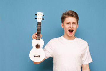 Emotional guy in a white T-shirt holds a ukulele in his hands, looking into the camera with an open mouth on a blue background. Isolated. Young man with ukulele on a colored background.