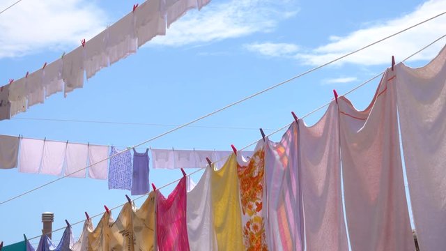 CLOSE UP: Warm summer air dries the freshly washed laundry hung out to dry on a sunny day in Greece. Colorful sheets, clothing and towels hang from the clotheslines under the clear blue summer sky.