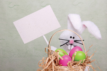 Easter bunny holding a blank card behind a wicker basket full of colorful Easter eggs