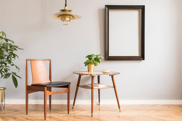 Stylish and design retro interior with design chair, gold lamp, small table with plant. Black mock up frame on the grey background wall. Minimalistic concept of sitting room with plants. Real photo. 