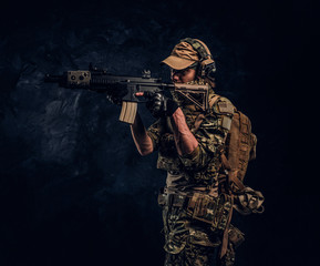 The elite unit, special forces soldier in camouflage uniform holding an assault rifle with a laser sight and aims at the target. Studio photo against a dark wall