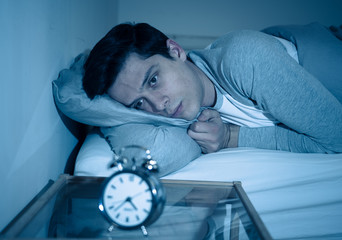 Portrait of young man in bed staring at alarm clock trying to sleep feeling stressed and sleepless