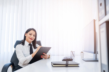 Satisfied young business woman writing on clipping board while sitting in her office.