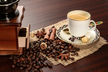 Cup of coffeee with saucer on a jute napkin. Vintage still life on wooden table with sugar, anise, cinnamon, grinder and dropped coffee beans