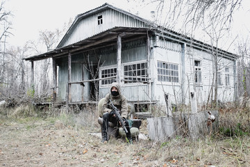 Stalker, a guy in uniform and with a machine gun in the yard of an old, abandoned house next to an empty parched well