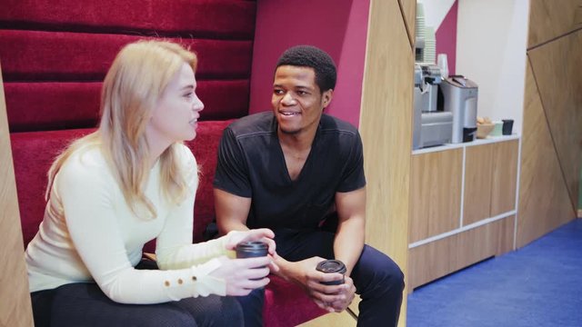Two diverse office workers spending coffee break together - sitting on sofa, talking and laughing