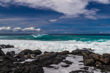 Shoreline of volcanic rocks on Hawaii's Big Island; foam and surf in foreground. Curling wave breaking offshore; in the background is the Pacific ocean, with blue skies and clouds overhead. 
