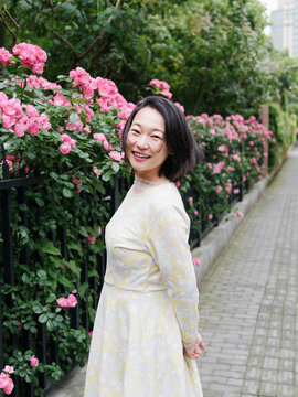 Outdoor Portrait Of Beautiful Middle Aged Chinese Woman In Yellow Dress Smiling Among Pink Rose Flowers Wall In Spring Garden, Happy Mature Woman Enjoy Her Leisure Time.