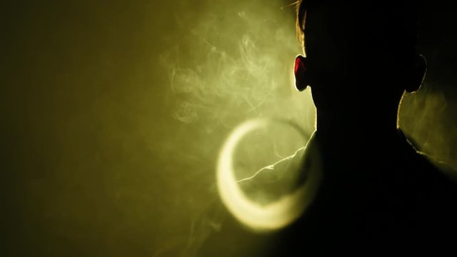 Silhouette of vaping man in the darkness. Boy blowing O's with vapor from e-cigarette. Vape tricks