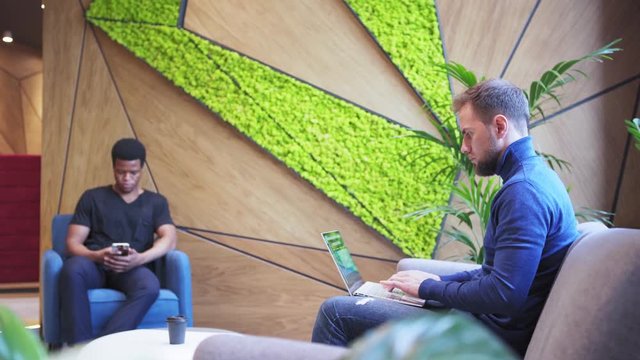 Side view of businessman in turtleneck sitting in armchair and working on laptop in office chill-out area; his colleague texting on cell phone in background