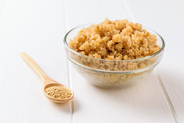 Wooden spoon with quinoa and a glass bowl with boiled quinoa on a white table.