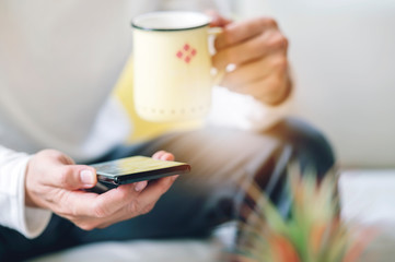 Cropped shot of man holding cup and using smartphone while relaxing at home.